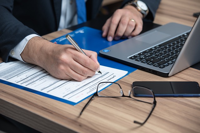 Persona in abito professionale che scrive su un documento, con laptop, occhiali e smartphone sulla scrivania, simbolo del calcolo della parcella legale.